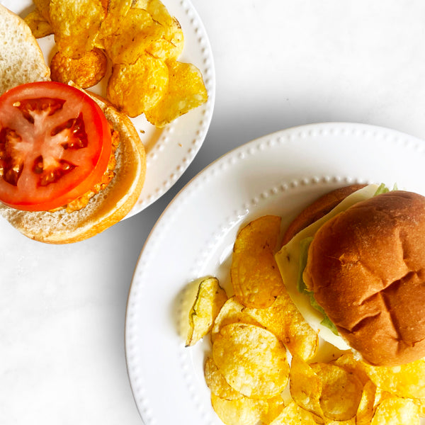 A top-down view of two plates, each with a turkey burger and a side of golden potato chips. One burger shows a slice of tomato, while the other is fully assembled with lettuce.