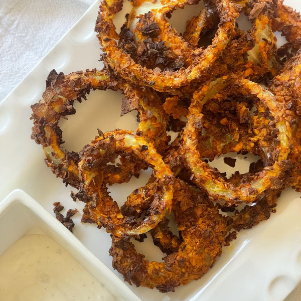 Crispy, golden-brown onion rings with a textured coating on a white rectangular dish, accompanied by a small container of dipping sauce.
