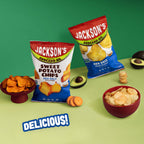 Two bags of Jackson's Avocado Oil potato chips are displayed on a light green surface against a solid green background, surrounded by food props including sliced avocados, bowls of chips, and a small stack of crackers. One bag is Sweet Potato Chips (Sea Salt) and the other is Regular Potato Chips (Sea Salt).