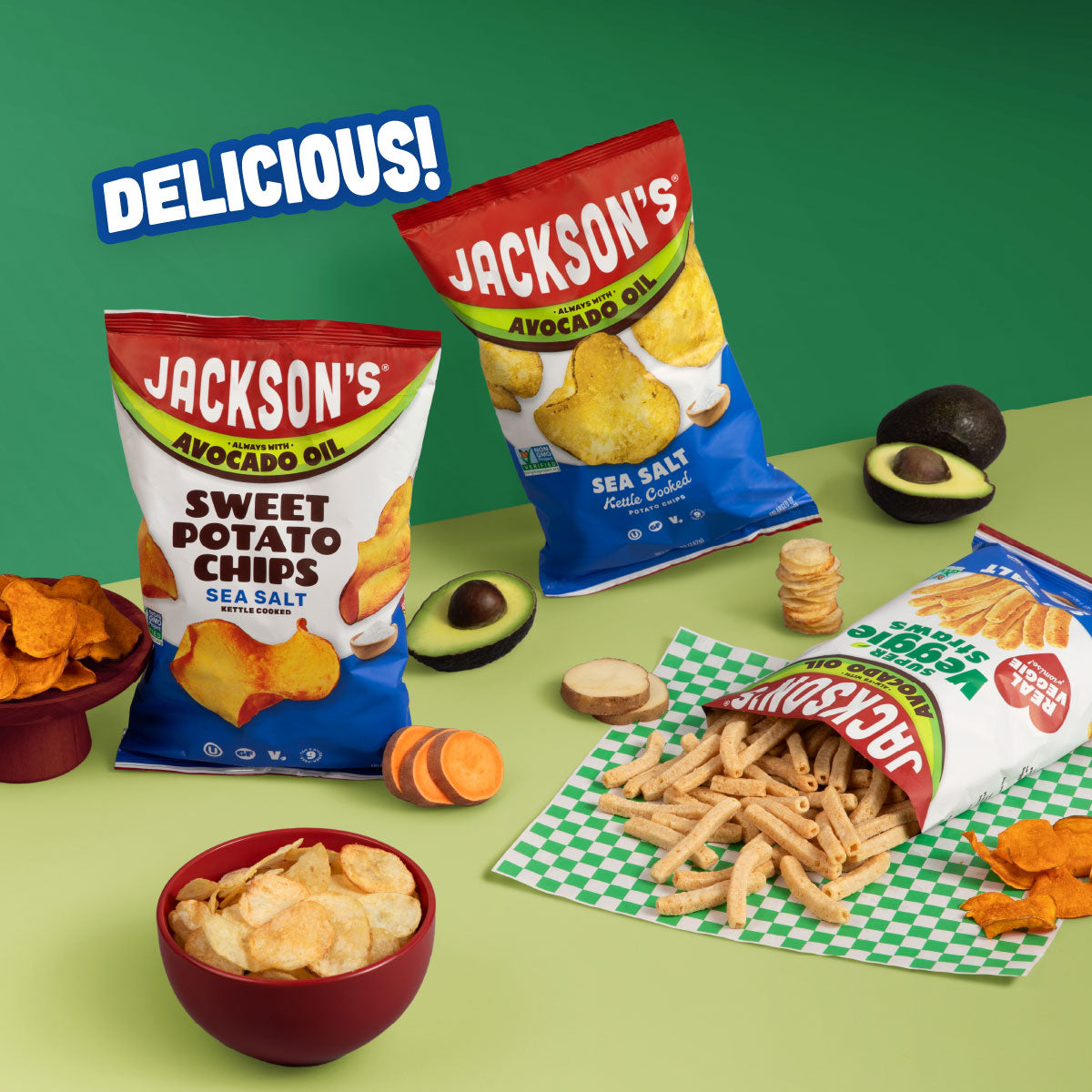 Three bags of Jackson's Avocado Oil snack chips arranged on a green tabletop with avocados, chips, and a bowl. The bags show Sweet Potato Chips Sea Salt, Potato Chips Sea Salt, and Super Veggie Straws.