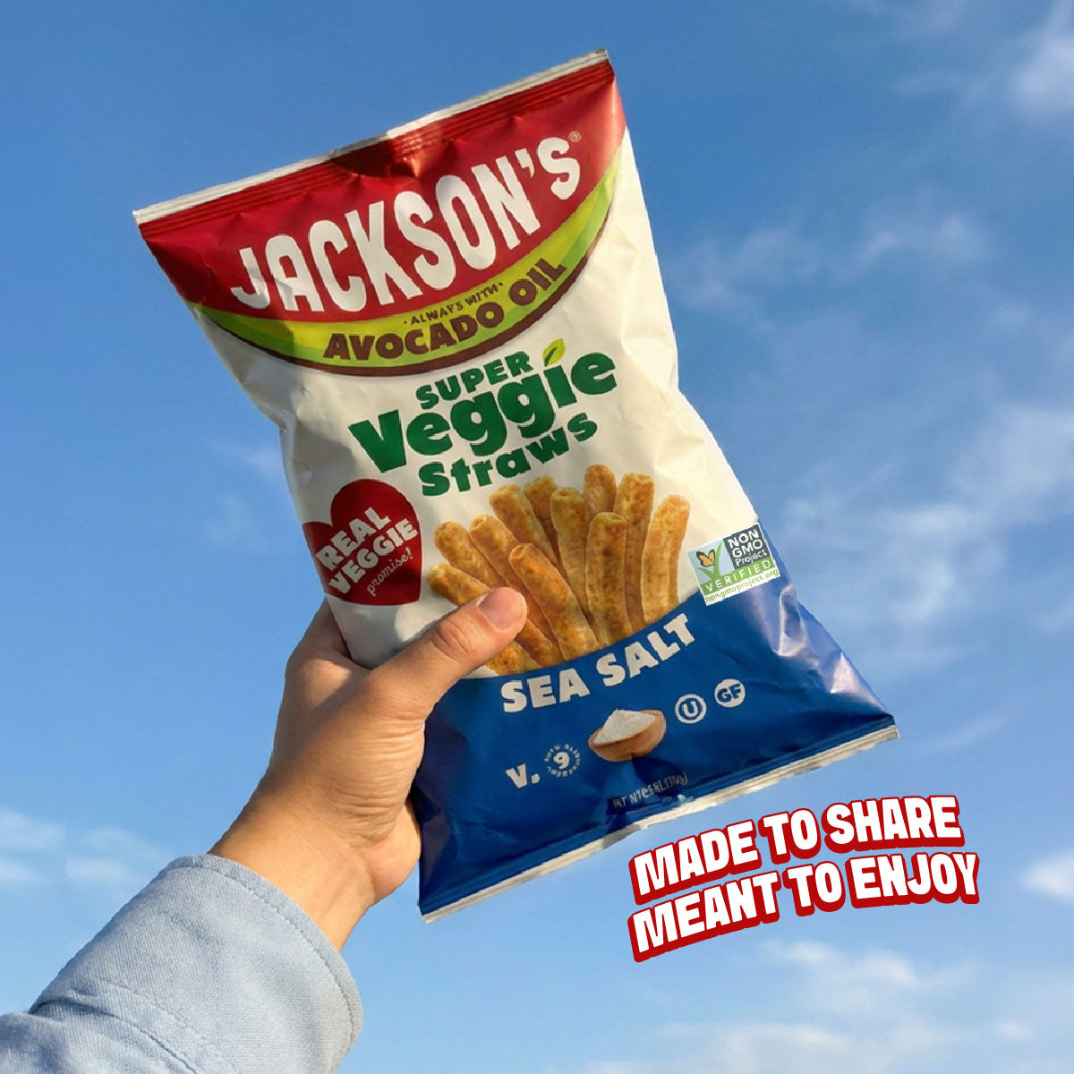 A hand holding a bag of Jackson's Super Veggie Straws Sea Salt against a blue sky background, with a red and white 'Made to Share, Meant to Enjoy' sticker overlay in the bottom right corner.