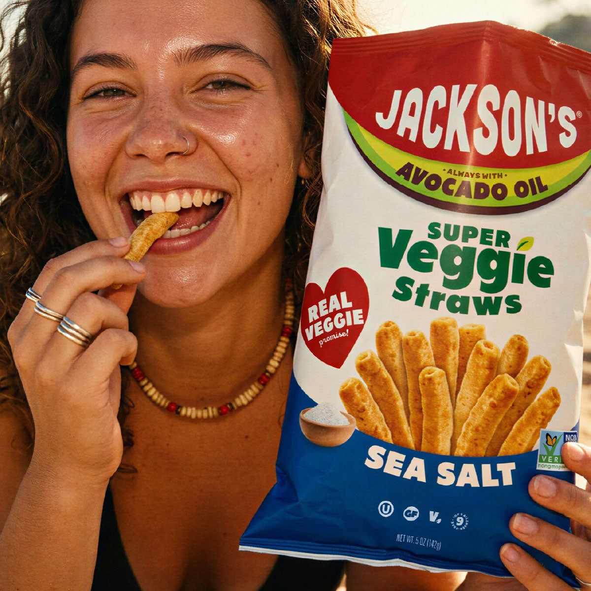 A woman smiling and eating a veggie stick while holding a bag of Jackson's Super Veggie Straws Sea Salt in one hand, with her face and hands visible in a close-up lifestyle shot.