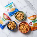 Three bags of Jackson's Super Veggie Sticks in different flavor variants are arranged on a white marble surface with four bowls of the golden-brown vegetable sticks.