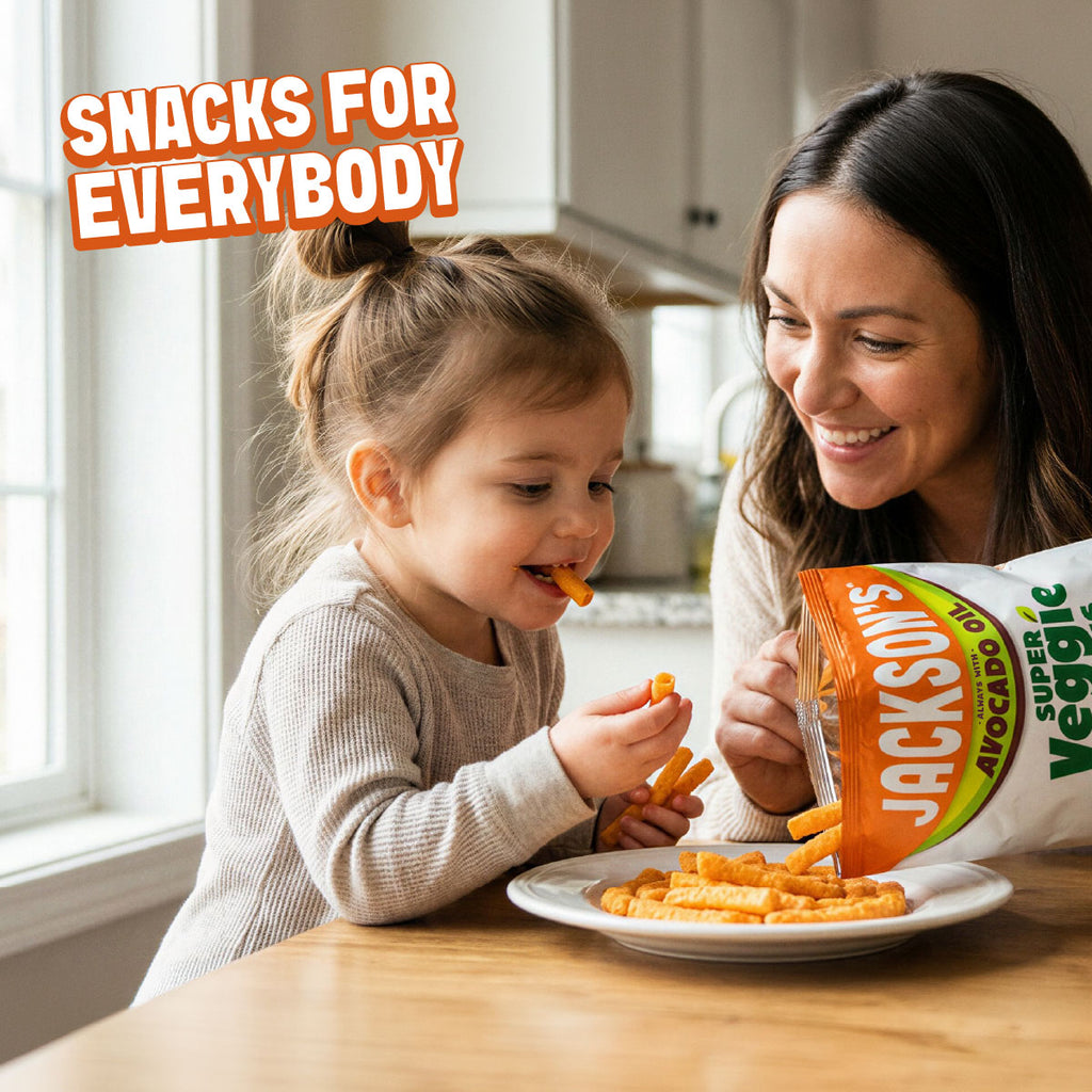 A woman and a young girl sit at a kitchen table smiling while eating orange snack sticks from a bag labeled Jackson's Super Veggie Avocado Chips. The text 'SNACKS FOR EVERYBODY' appears in the top left corner.