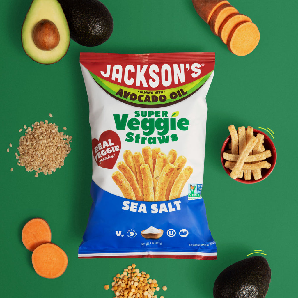 A bag of Jackson's Super Veggie Straws Sea Salt chips is centered on a bright green background, surrounded by food props including sliced avocados, sweet potatoes, and a small bowl of the chips.