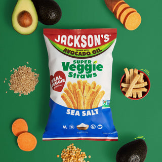 A bag of Jackson's Super Veggie Straws Sea Salt chips is centered on a bright green background, surrounded by food props including sliced avocados, sweet potatoes, and a small bowl of the chips.