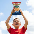 A young boy in a red t-shirt holds a bag of Jackson's Sea Salt Veggie Straws above his head against a blue sky with clouds.