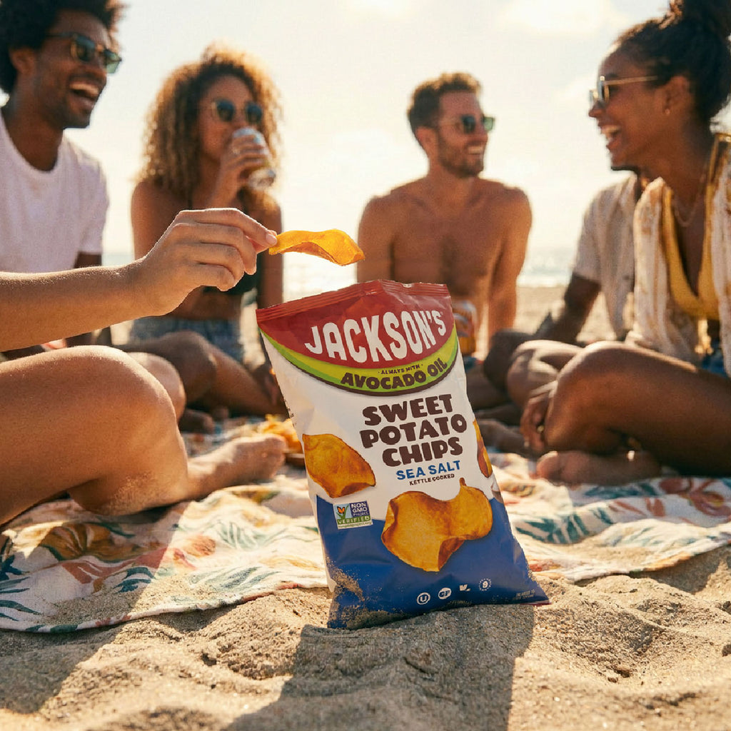 A bag of Jackson's Sweet Potato Chips Sea Salt is placed on a sandy beach, with a hand reaching in to grab a chip. In the background, a group of friends relaxes on a blanket by the ocean during golden hour.