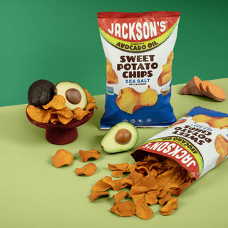 Two bags of Jackson's Sweet Potato Chips are displayed on a light green surface with a dark green background. One bag is standing upright on a small red pedestal, and the other lies on its side with chips spilled out.