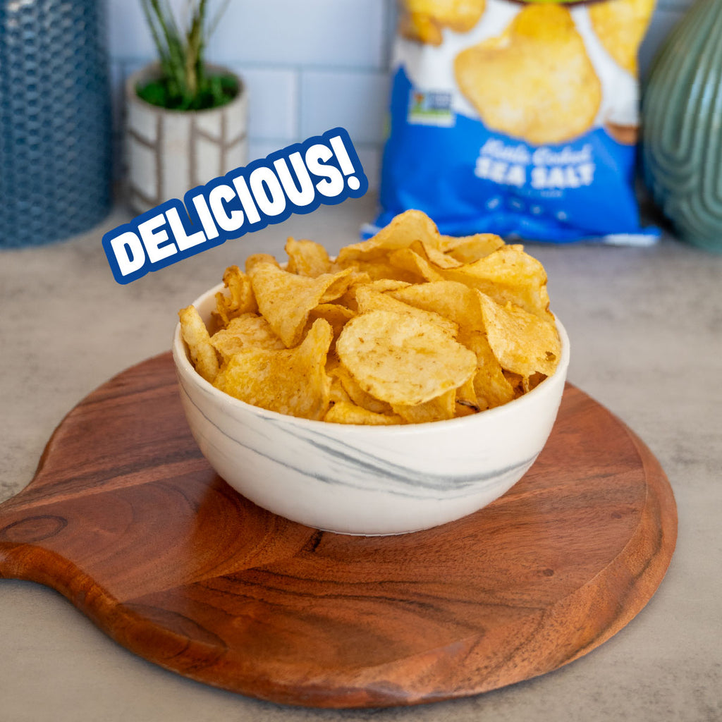 A white marble-patterned bowl filled with golden, crispy potato chips sits on a round wooden serving board. In the background, a blue bag of Sea Salt potato chips is visible, with a blue 'DELICIOUS!' text overlay on the left.