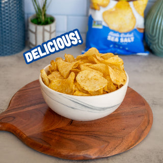 A white marble-patterned bowl filled with golden, crispy potato chips sits on a round wooden serving board. In the background, a blue bag of Sea Salt potato chips is visible, with a blue 'DELICIOUS!' text overlay on the left.