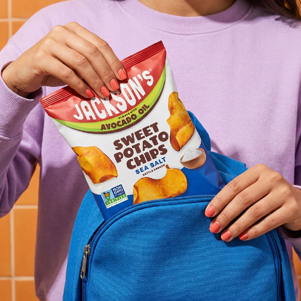 A person in a lavender sweatshirt holds a bag of Jackson's Sweet Potato Chips in front of a bright blue backpack, with orange-tiled wall visible in the background.