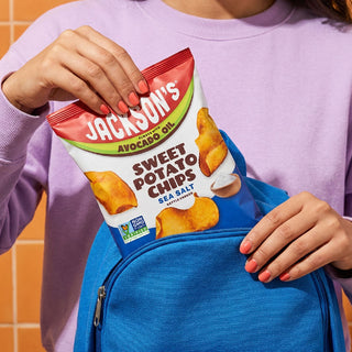 A person in a lavender sweatshirt holds a bag of Jackson's Sweet Potato Chips in front of a bright blue backpack, with orange-tiled wall visible in the background.