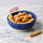 A blue ceramic bowl filled with golden-brown crunchy stick-shaped snacks, placed on a white marble surface with one snack resting outside the bowl. A red and white 'CRUNCHY!' text badge appears near the top left.