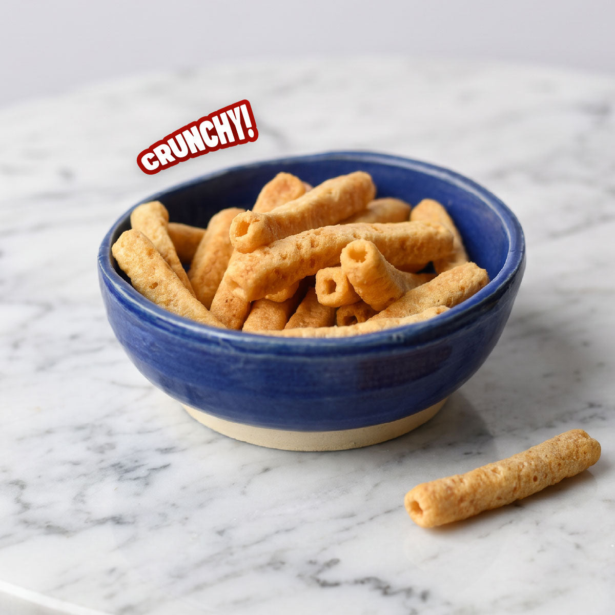 A blue ceramic bowl filled with golden-brown crunchy stick-shaped snacks, placed on a white marble surface with one snack resting outside the bowl. A red and white 'CRUNCHY!' text badge appears near the top left.
