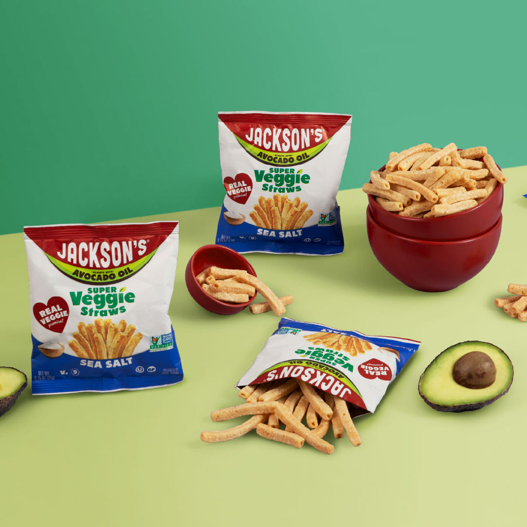 Three bags of Jackson's Avocado Oil Super Veggie Straws Sea Salt are arranged on a light green surface with bowls of the golden fried straws and avocado props against a solid green background.