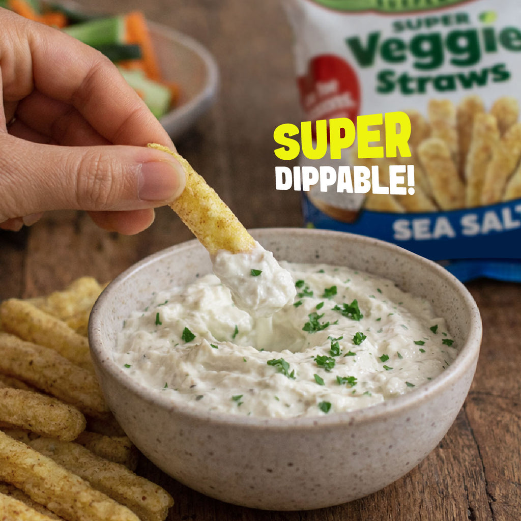 A hand holds a super veggie straw above a bowl of creamy dip with green herb garnish, with Veggie Straws packaging and a bag of sea salt sticks visible in the background on a wooden table. Large text reads 'SUPER DIPPABLE!'.