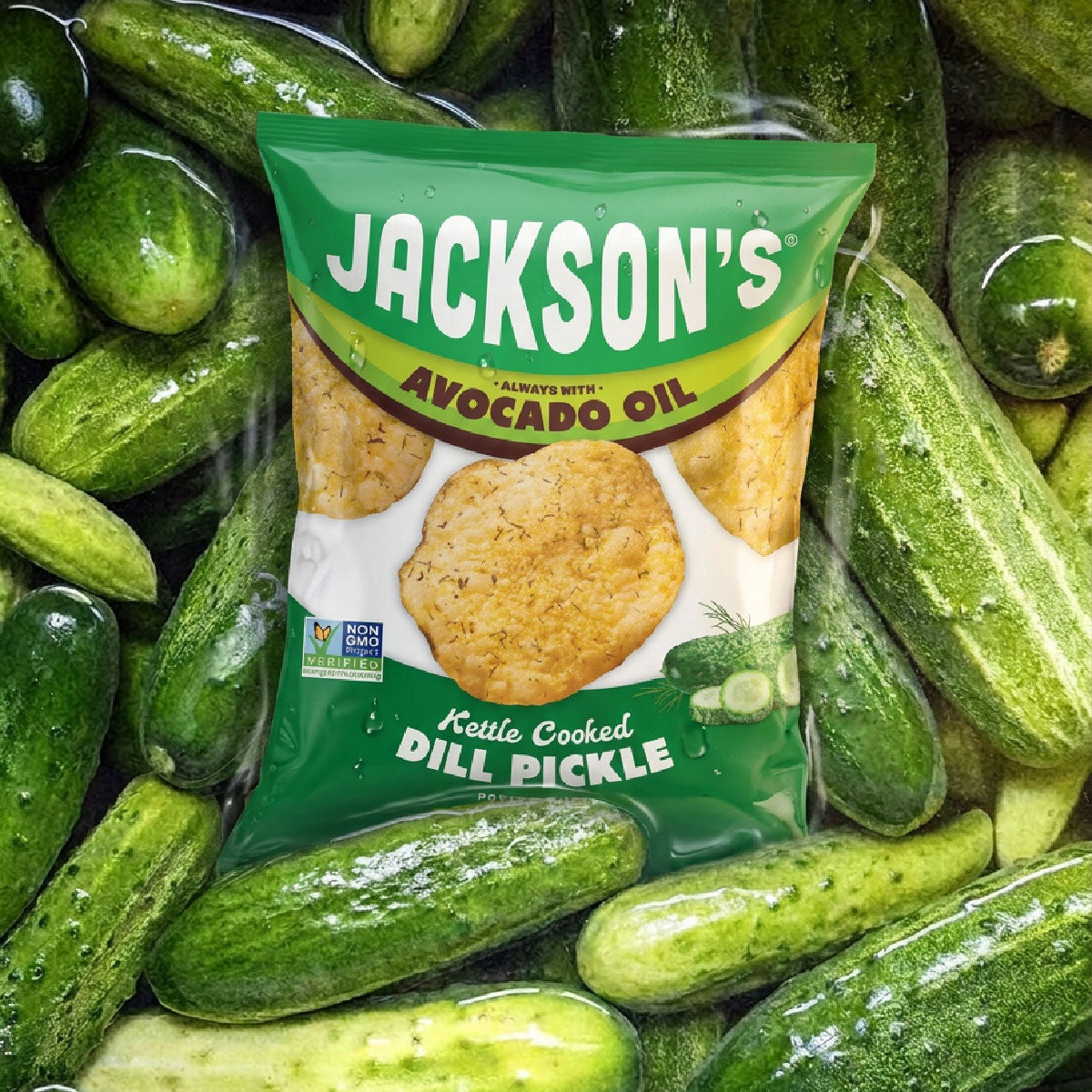 A bag of Jackson's Kettle Cooked Dill Pickle chips surrounded by fresh green cucumbers. The bag is green with white and yellow text, showing avocado oil and pickle imagery.