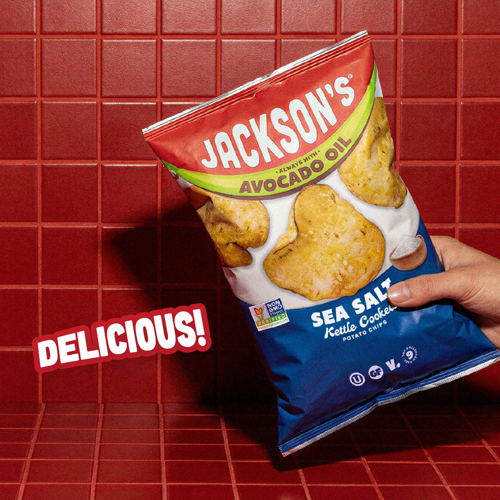 A hand holding a bag of Jackson's Sea Salt Keto Cookie Potato Chips against a red tiled background, with a 'DELICIOUS!' text overlay.