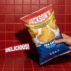 A hand holding a bag of Jackson's Sea Salt Keto Cookie Potato Chips against a red tiled background, with a 'DELICIOUS!' text overlay.