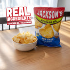 A bag of Jackson's Sea Salt Kettle Cooked Potato Chips sits on a wooden table next to a white bowl filled with chips. A graphic overlay in the top left reads 'REAL INGREDIENTS +AVOCADO OIL + SEASONING'.