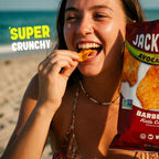 A woman on a beach holds a bag of Jackson 'Barbecue Kettle Crisp' potato chips, eating a chip while smiling. The bag is red with white and green text, and the background shows sand and the ocean.