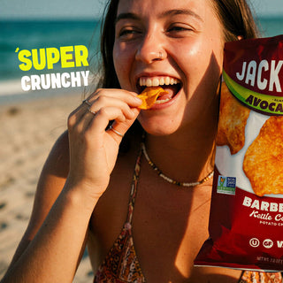A woman on a beach holds a bag of Jackson 'Barbecue Kettle Crisp' potato chips, eating a chip while smiling. The bag is red with white and green text, and the background shows sand and the ocean.