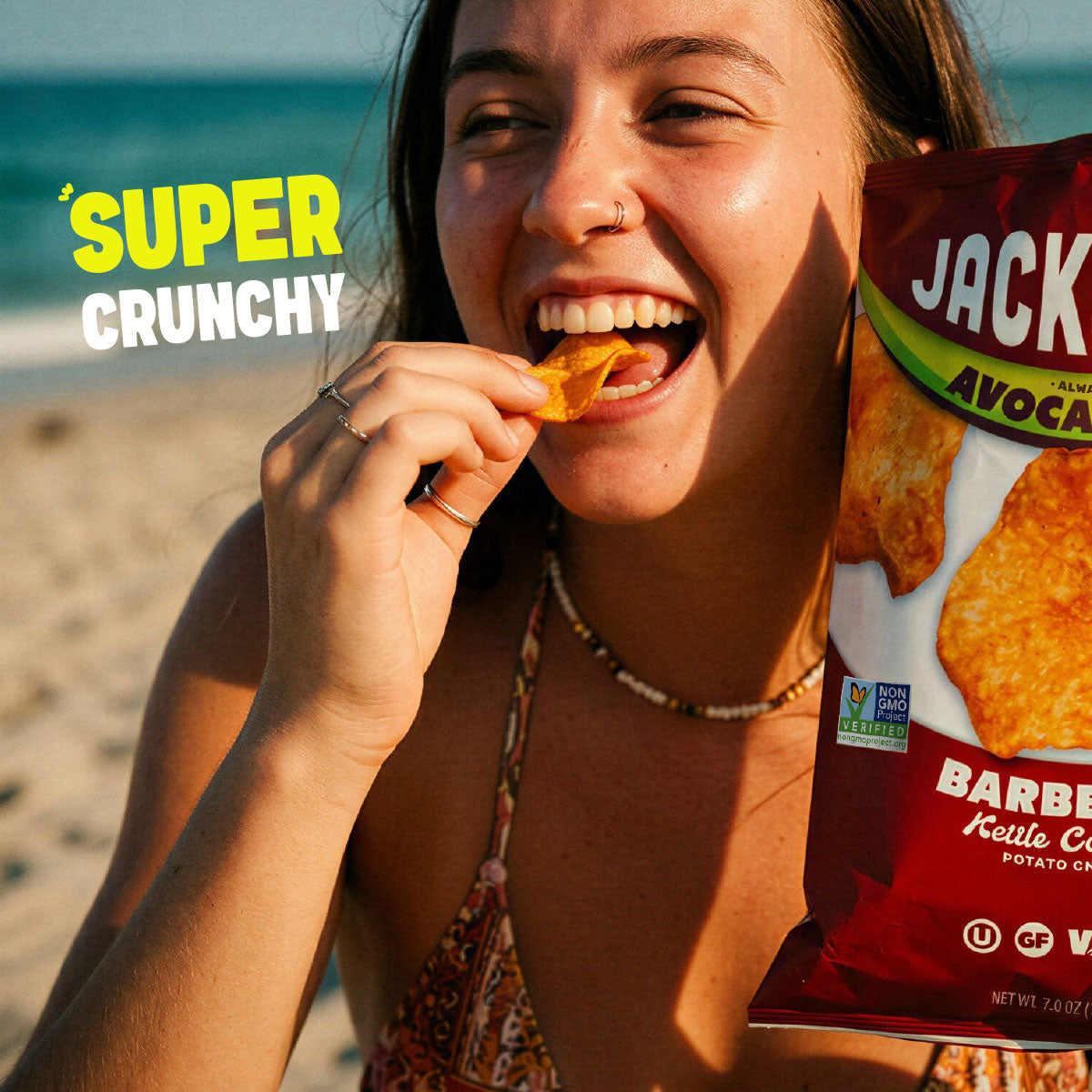 A woman on a beach holds a bag of Jackson 'Barbecue Kettle Crisp' potato chips, eating a chip while smiling. The bag is red with white and green text, and the background shows sand and the ocean.