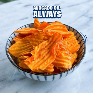 A bowl of golden-brown potato chips filled with orange avocado oil, set on a white marble surface with a text overlay that reads 'AVOCADO OIL ALWAYS'.