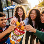 Four smiling young people reaching into a bag of Jackson's Avocado Oil Sweet Potato Chips Sea Salt outdoors near a bus stop. The bag is blue, red, and white, and the scene is bright with warm sunlight.