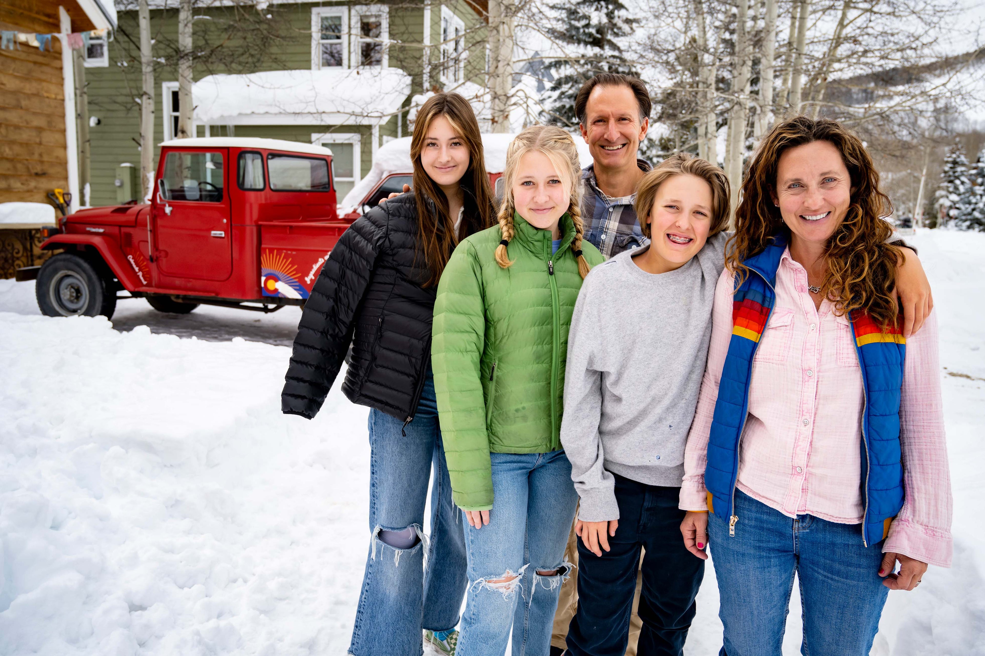 A family of five posing together outdoors in the snow in front of a house and a red pickup truck. The group is dressed in winter clothing, including puffer jackets, a gray hoodie, and a pink shirt with a blue and rainbow vest.