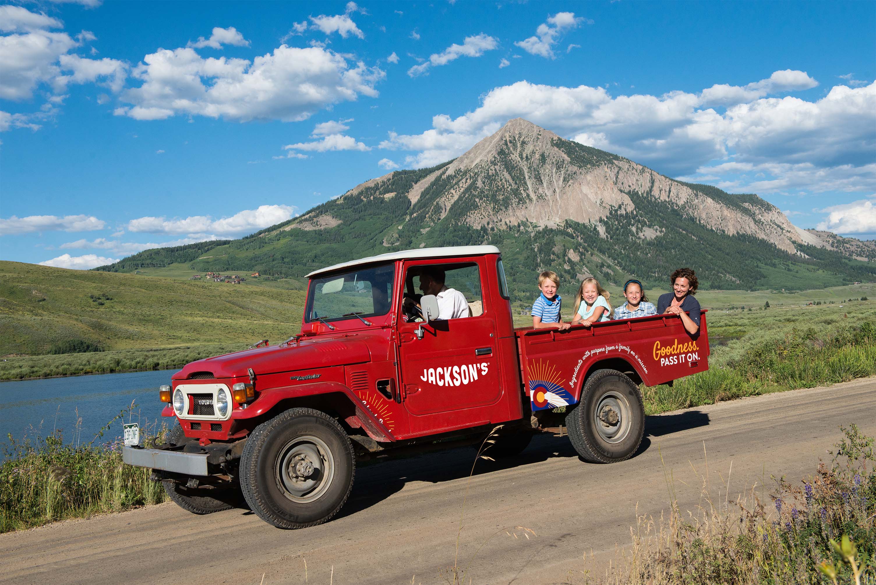 A red vintage pickup truck with four children in the open back drives along a road with a mountain and lake in the background. The side of the truck reads 'JACKSON'